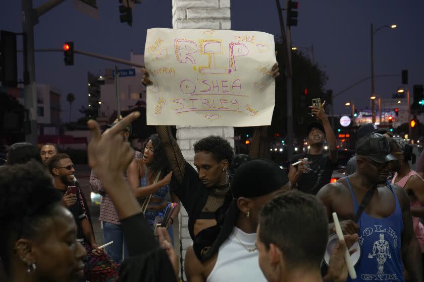 Someone standing at the center of a gas station holding up a sign that reads "RIP O'Shae Sibley" as other strut past.