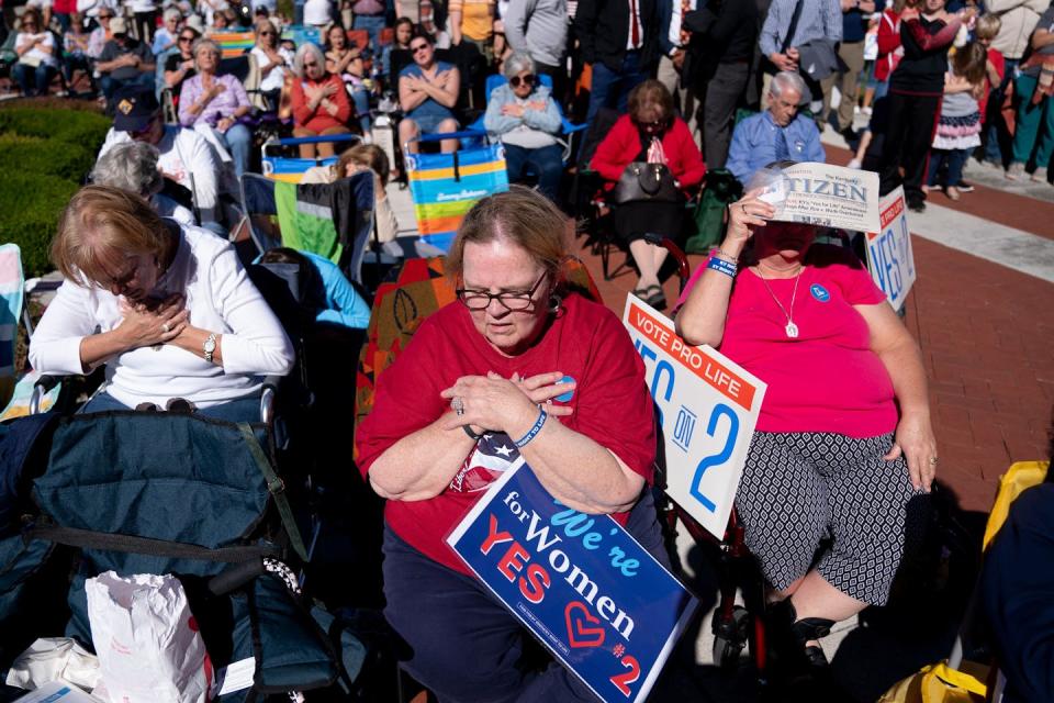 People sit in folding chairs in a row and cross their arms and look down. One holds a poster that says 'We're women for yes.'