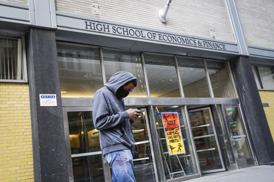 FILE - In this March 16, 2020, file photo, a pedestrian wears a face mask while standing outside the High School of Economics & Finance, closed due to coronavirus concerns, in New York. Most New York City students will return to their physical schools two or three days a week and learn online the rest of the time under a plan announced Wednesday, July 8, 2020, by Mayor Bill de Blasio, who said schools can't accommodate all their students at any one time and maintain safe social distancing. (AP Photo/John Minchillo, File)