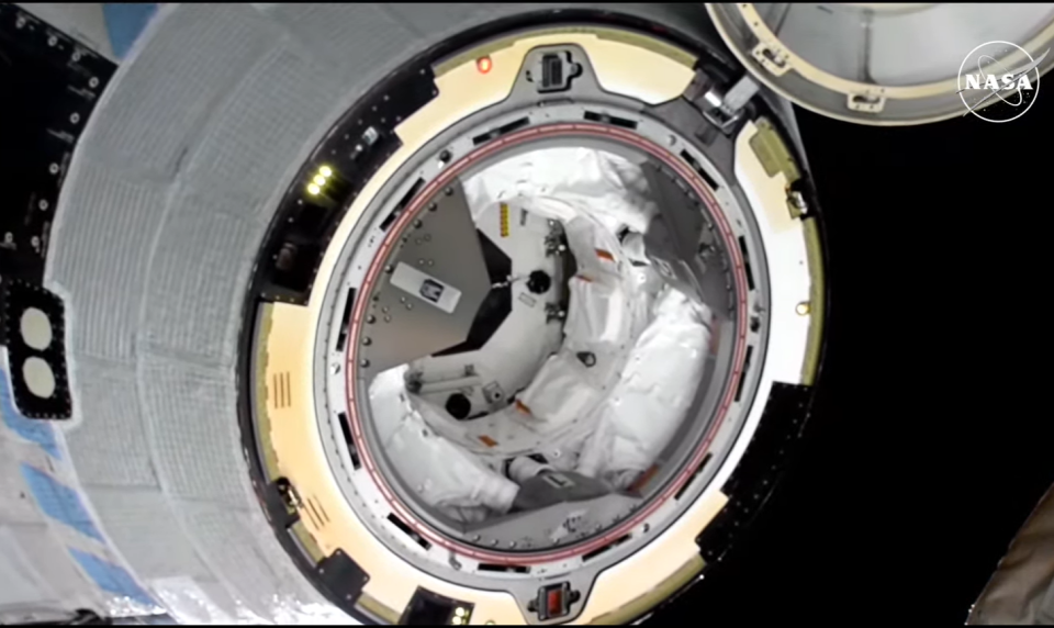 The Starliner capsule is seen in space after undocking from the ISS. It is a white capsule with a hatch open and a sealed area within.