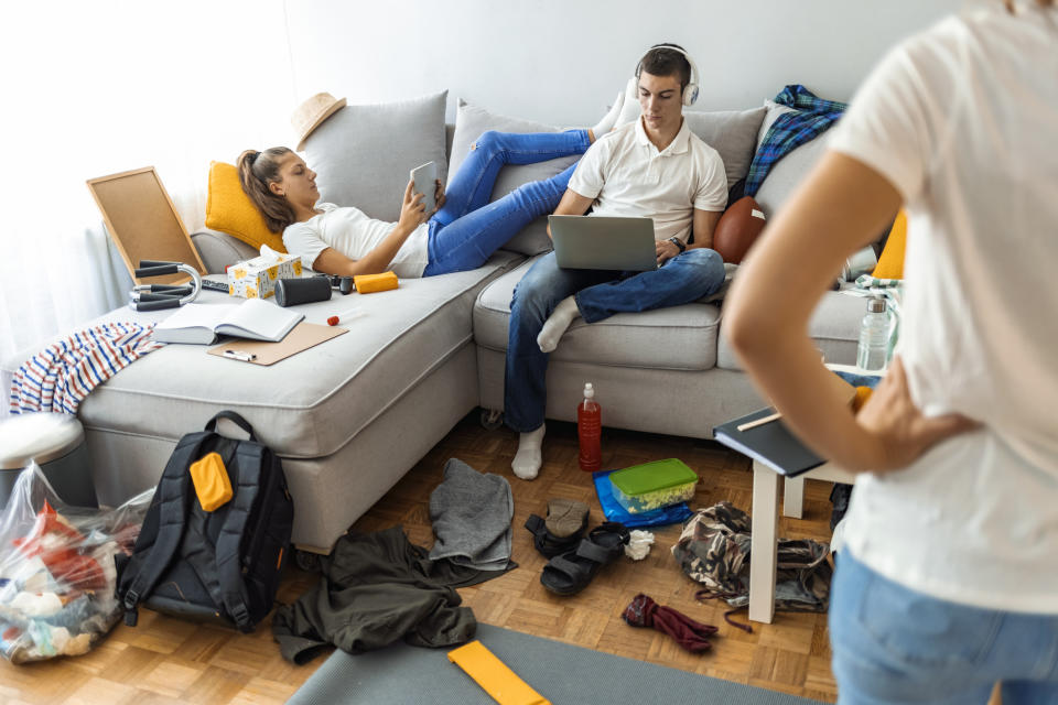 Two teenagers using electronic devices on a sofa in a messy living room as an adult stands in the foreground