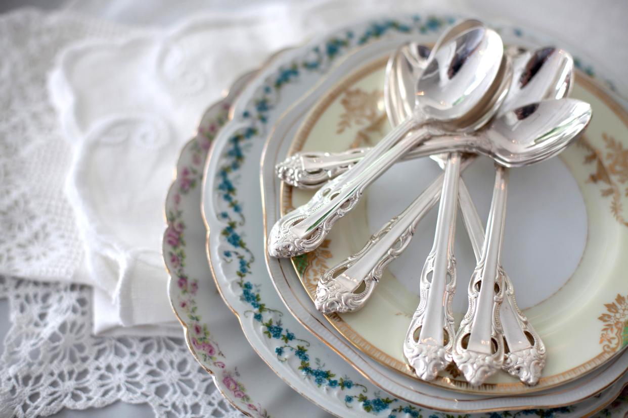 a stack of vintage floral plates with silver teaspoons on a table near lace doilies