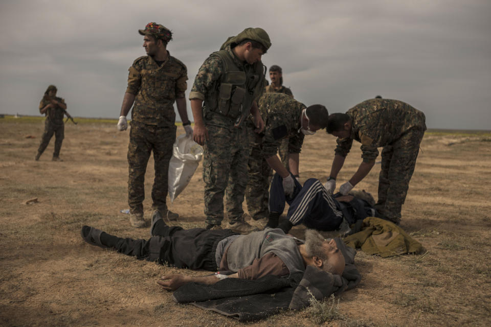 In this Sunday, March 10, 2019 photo, U.S.-backed Syrian forces stand guard as civilians wounded by airstrikes, during an offensive on the last area held by Islamic State group extremists, are treated at a reception area for evacuees, in Syria's eastern Deir el-Zour province near the Iraqi border, outside of Baghouz, Syria.