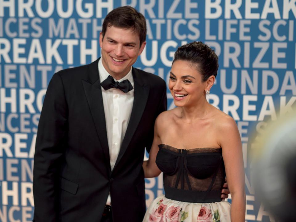 Ashton Kutcher with his arm around Mila Kunis on the red carpet in front of a blue step-and-repeat.