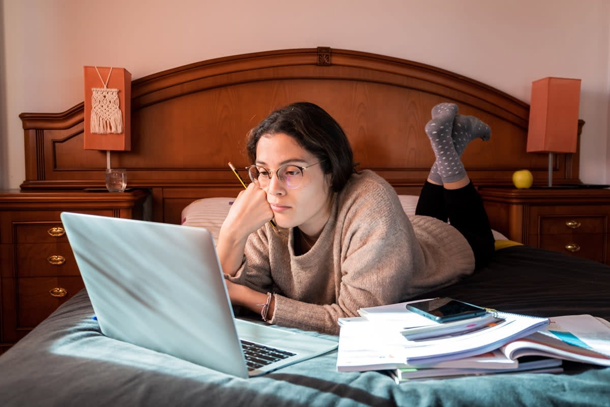 A woman does school work on her laptop while laying on her bed.