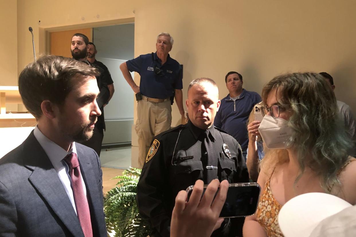 At left man in a suit talks with a young person on the right, while other people, including a police officer, stand around.