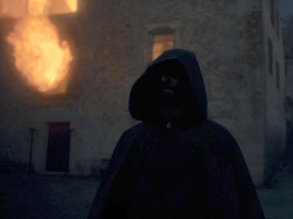 A hooded figure walks away from a fire in a building.