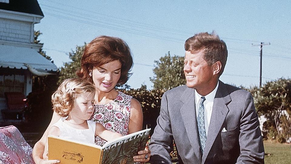 Jacqueline Kennedy and her husband Senator John F. Kennedy sit on a lounge chair at their summer home. Jacqueline reads to her daughter Caroline from a book.