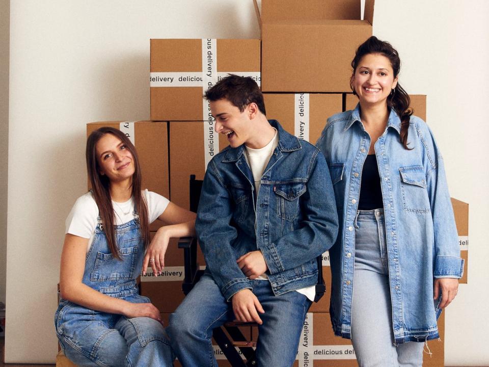 Three people wearing denim sit among a stack of brown boxes for a photo shoot.