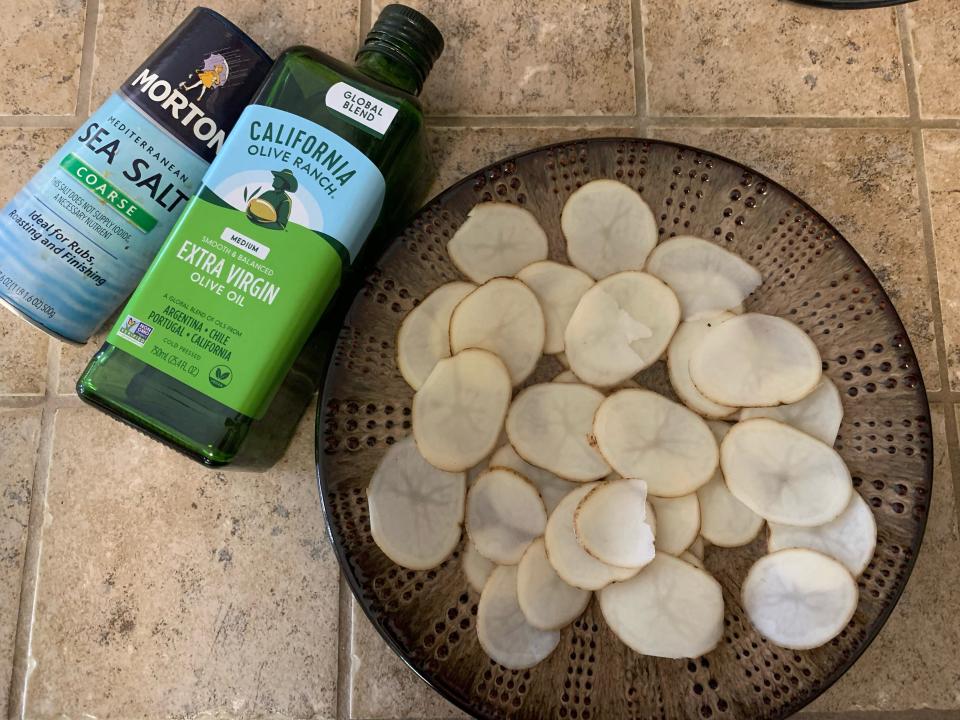 container of sea salt and bottle of olive oil beside a plate of sliced potato chips