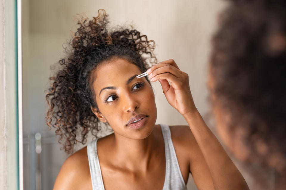 African american woman plucking eyebrows with tweezers while standing in front of the mirror. Closeup of young woman in pajamas removing facial hair in bathroom. Girl getting her eyebrows shaped during her morning beauty routine.