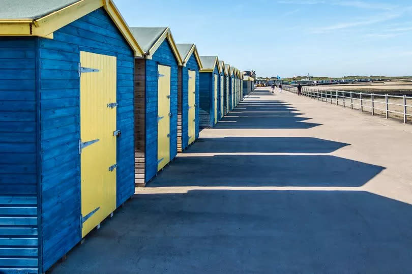 Blue and yellow wooden beach huts casting long shadows in the sunshine along the promenade at the coast of Minnis Bay, Kent, UK