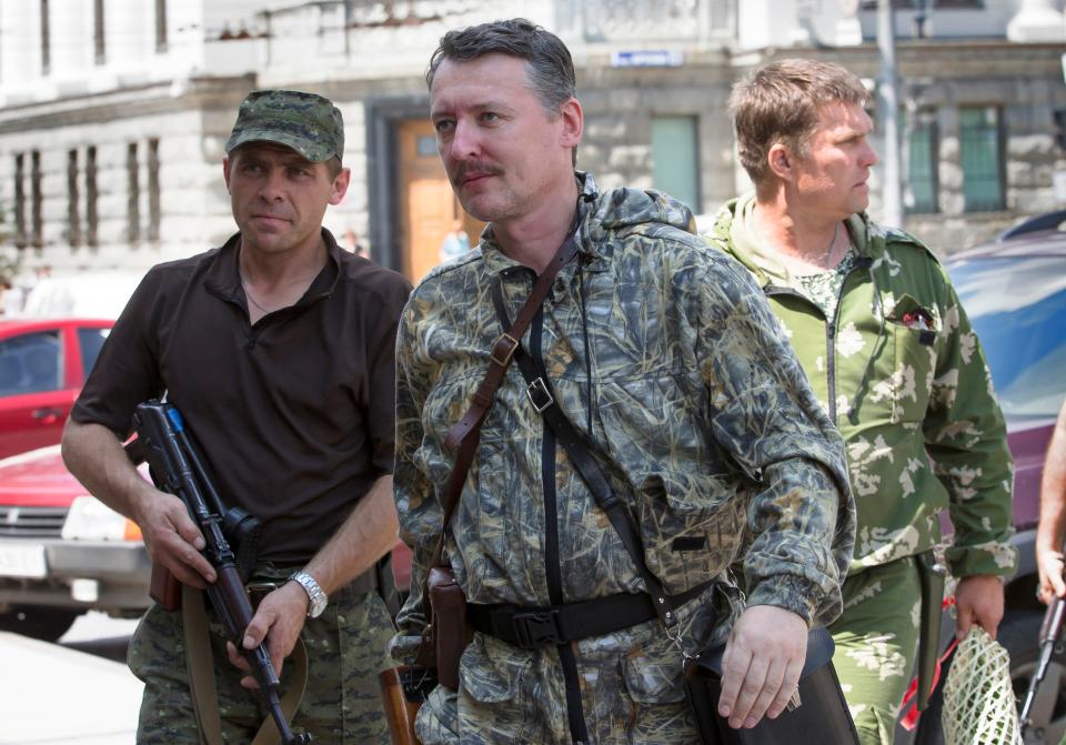 A man in military dress walks forward as two men stand nearby.