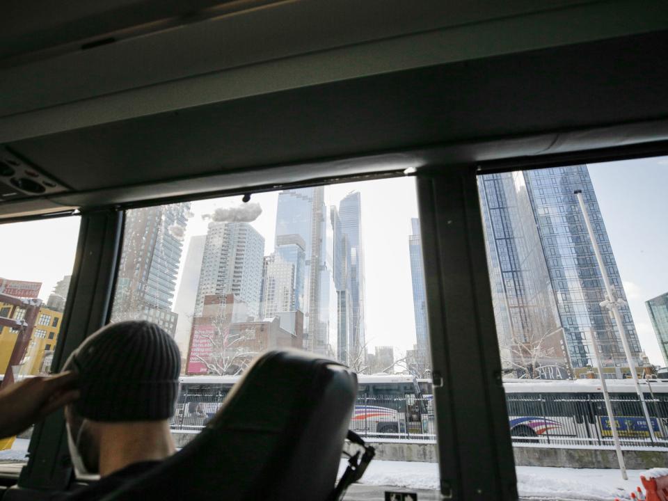 A view of tall buildings through bus windows. Someone wearing a beanie sits in the lower corner of the frame.