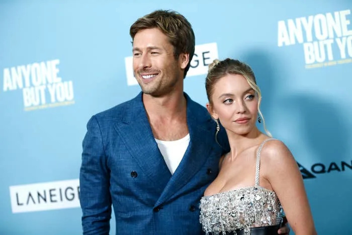 Glen Powell and Sydney Sweeney pose together at the "Anyone But You" event. Glen wears a double-breasted blazer; Sydney wears a bejeweled, glamorous dress