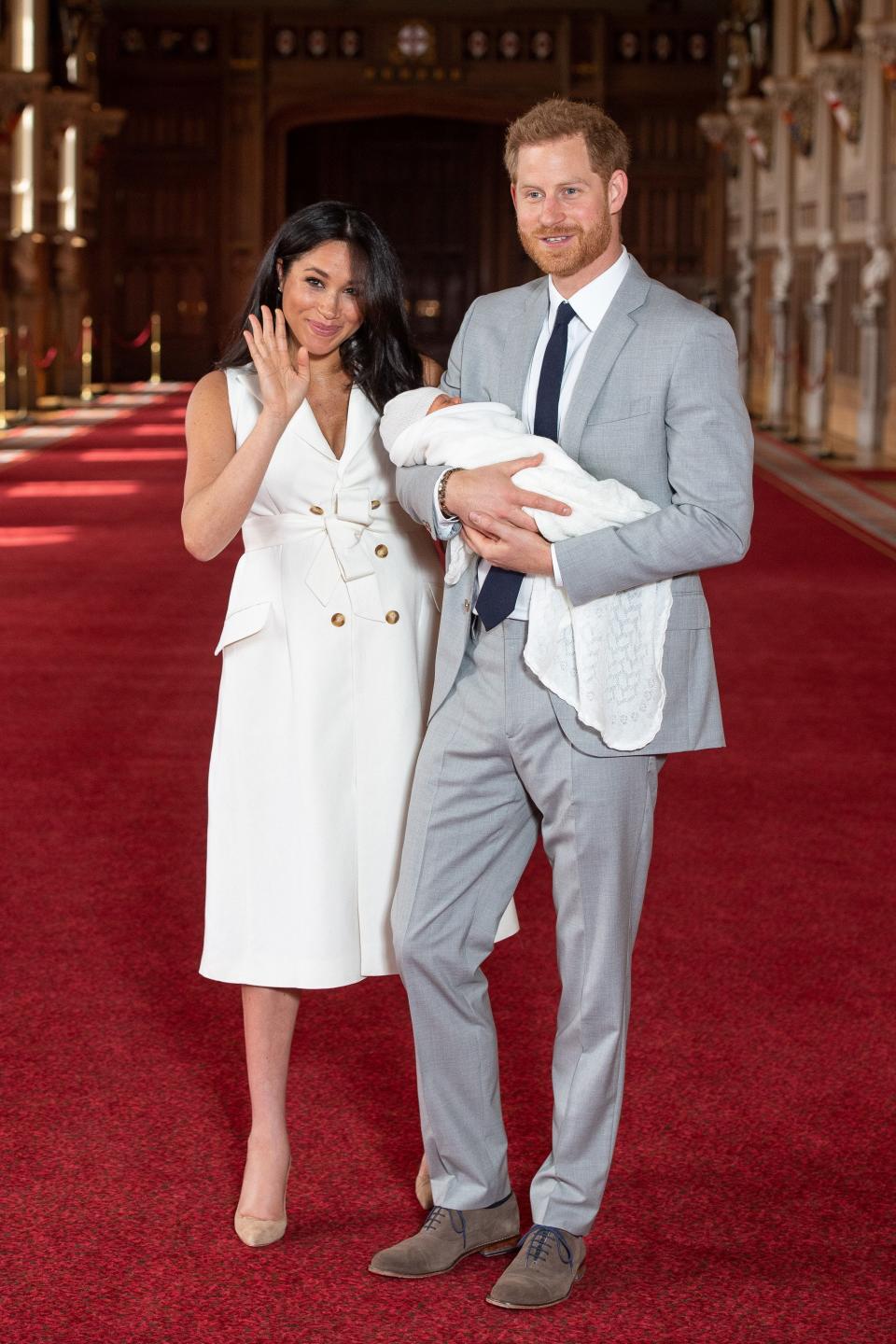 Meghan in a white button decorated tea length dress and Harry in a light grey suit and navy tie holding Archie.