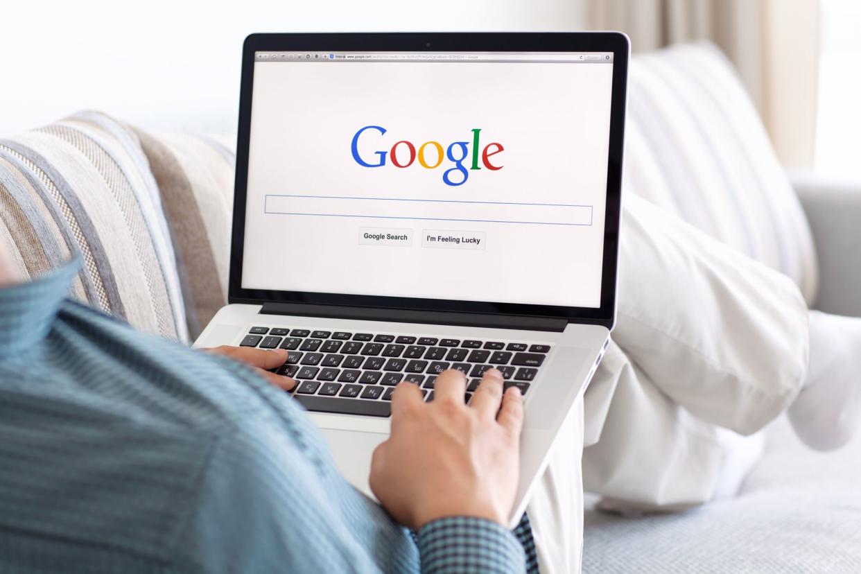 Google homepage on the Safari browser on the screen of a MacBook, man with back towards the camera, laying on a couch with laptop on his knees, blurred background