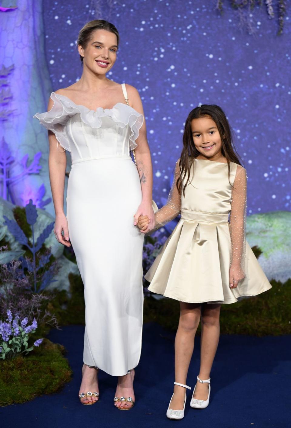 helen flanagan, a woman wearing a white dress, holding hands with a child, also in a white dress, smiling and standing in front of a purple background at the wish movie premiere in 2023