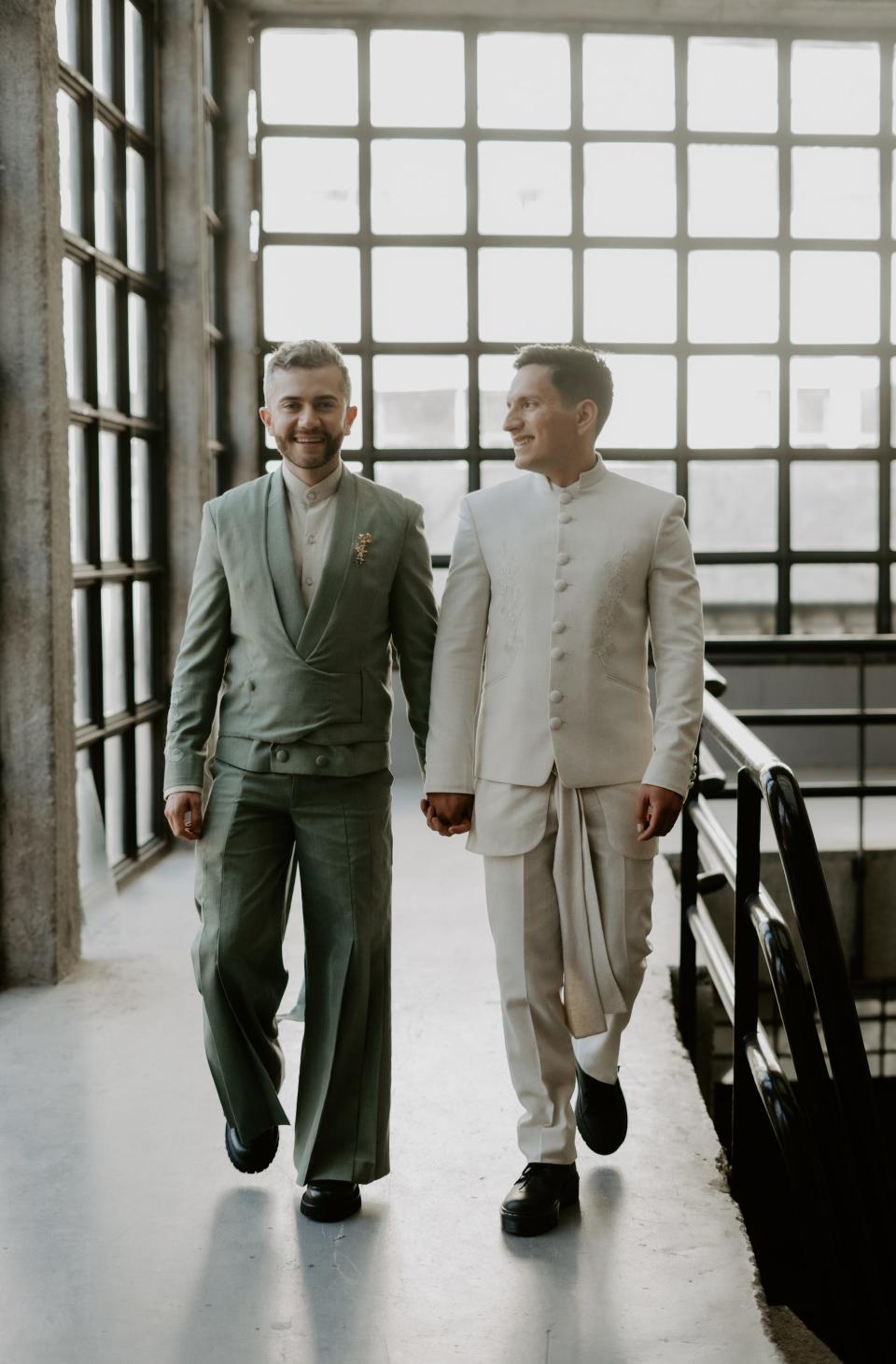 Two grooms hold hands as they walk through a room full of windows.