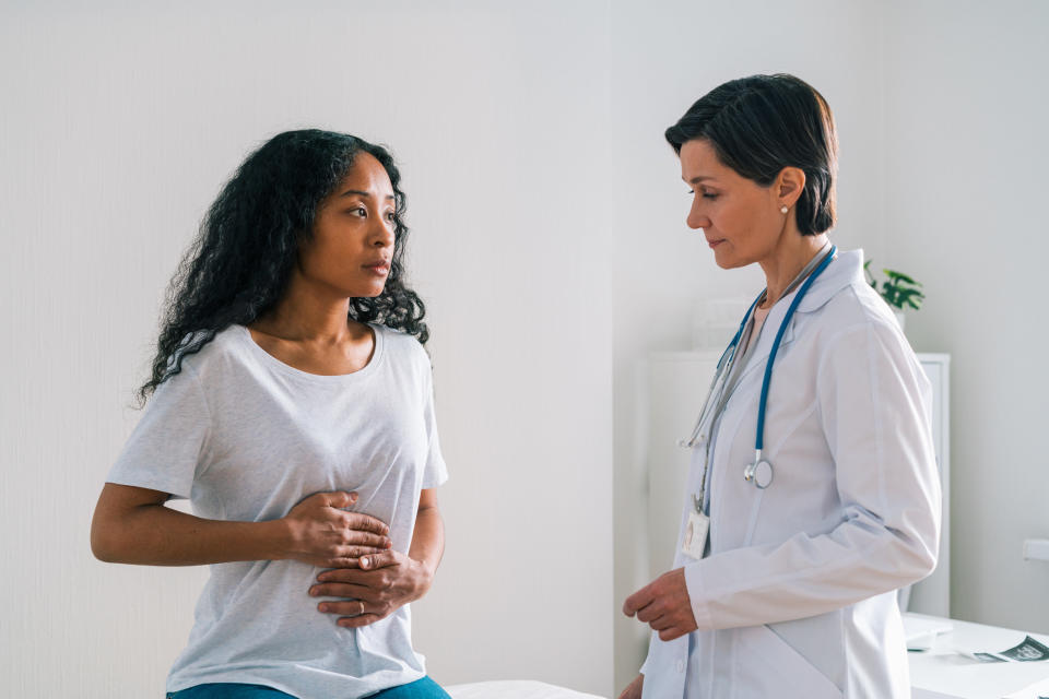 A patient holds their abdomen as they sit on the table in a clinic room as a doctor looks on