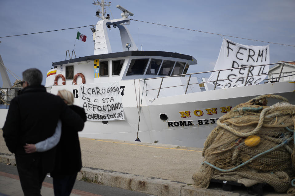 People walk past a fishing boat with banners against the gasoline price increase, in the Roman port of Fiumicino, Friday, March 11, 2022. Nowhere more than in Italy, the European Union’s third-largest economy, is dependence on Russian energy taking a higher toll on industry. (AP Photo/Andrew Medichini)