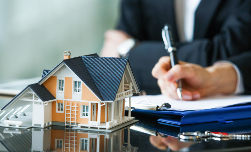 A person filling out an form with a small house sitting on a desk.