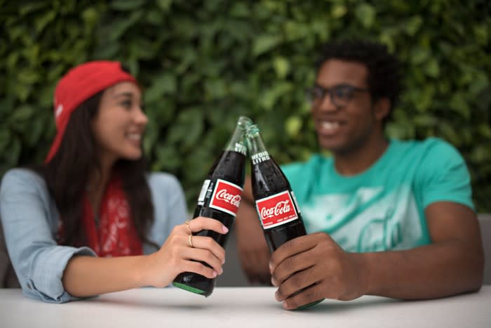 A man and woman clinking their Coca-Cola bottles together while seated and talking outside.