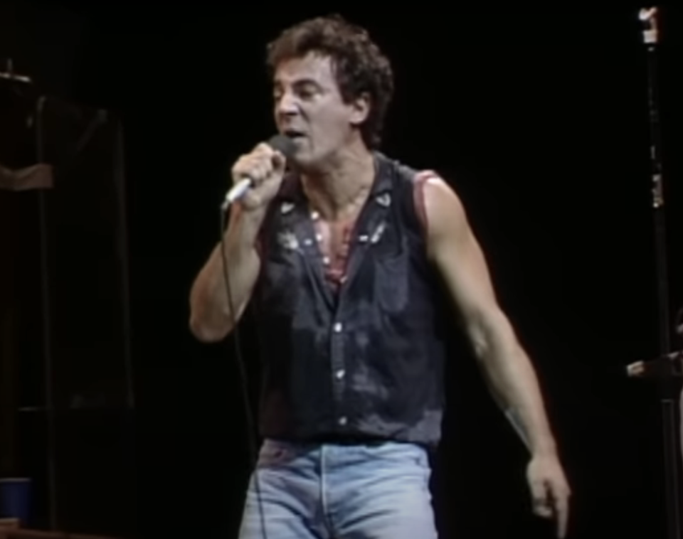 Bruce Springsteen on stage singing into microphone wearing a sleeveless top and necklace
