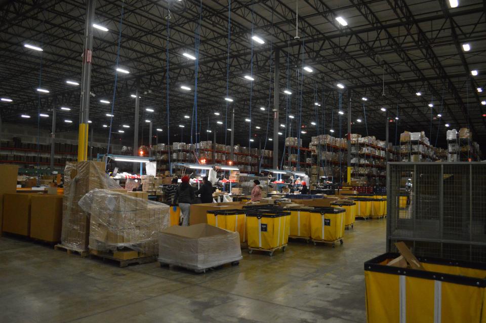 Bright yellow rolling bins sit in front of brightly-lit work stations in a large warehouse