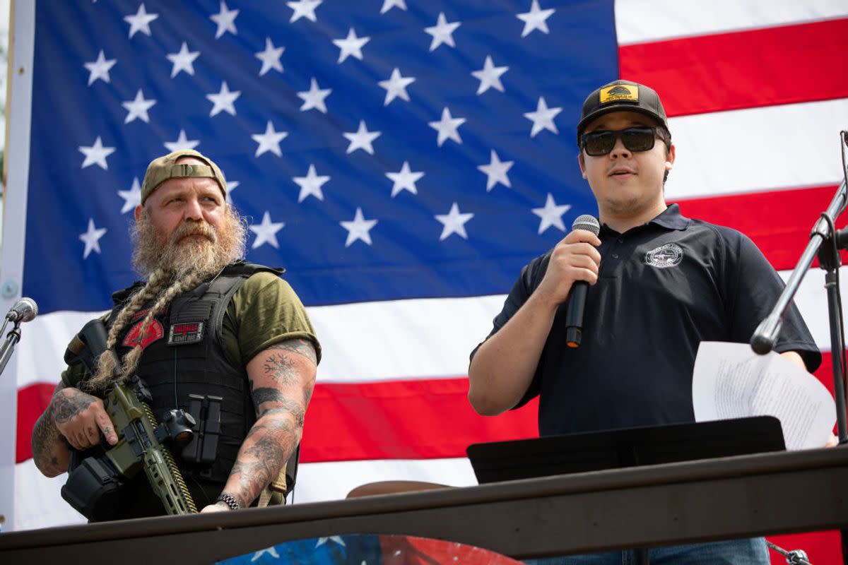 A white man holding a microphone stands next to another white man with a large gun strapped to him. 