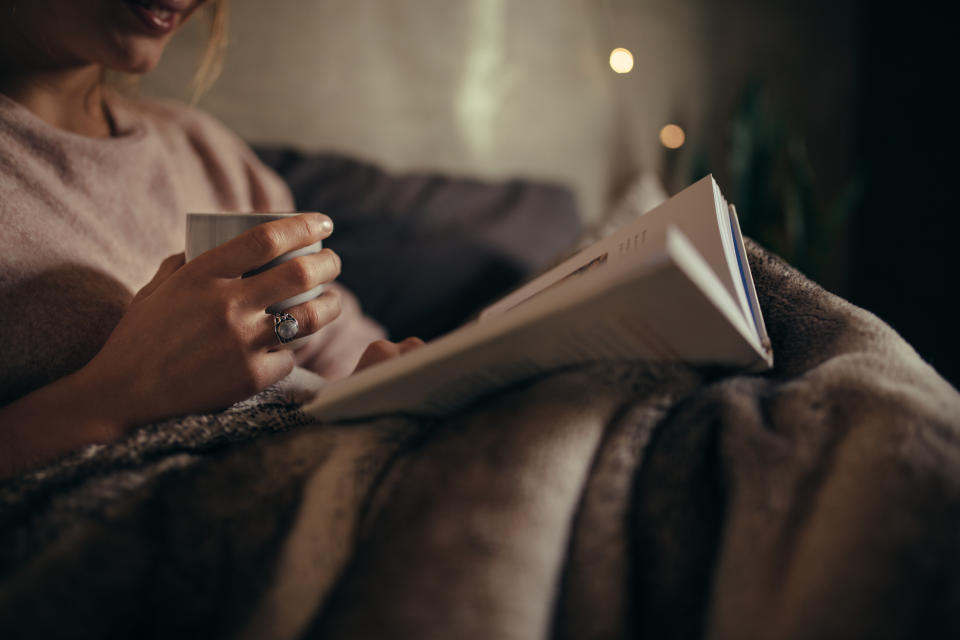 A photo of a woman in bed reading at while holding a cup of a drink. (Photo via Getty Images)