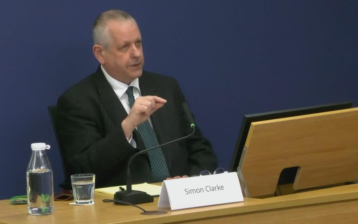 Simon Clarke gestures with his spectacles, seated at a desk bearing his name