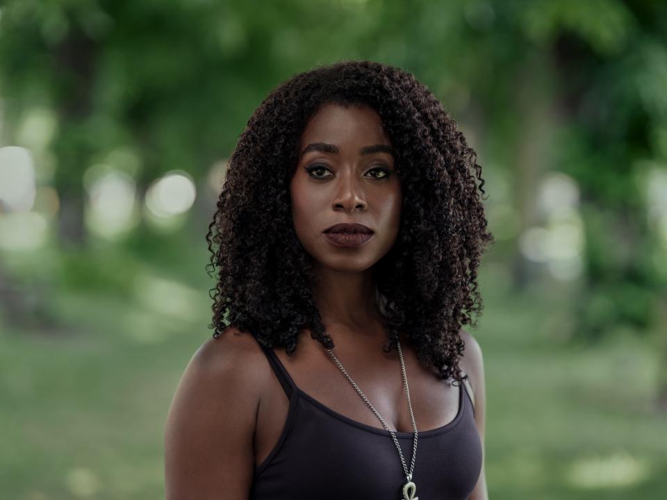 kirby howell-baptiste as death in the sandman. she's standing in a green space wearing a black tank top and golden necklace with an ankh symbol pendant, and her hair is worn down and curly