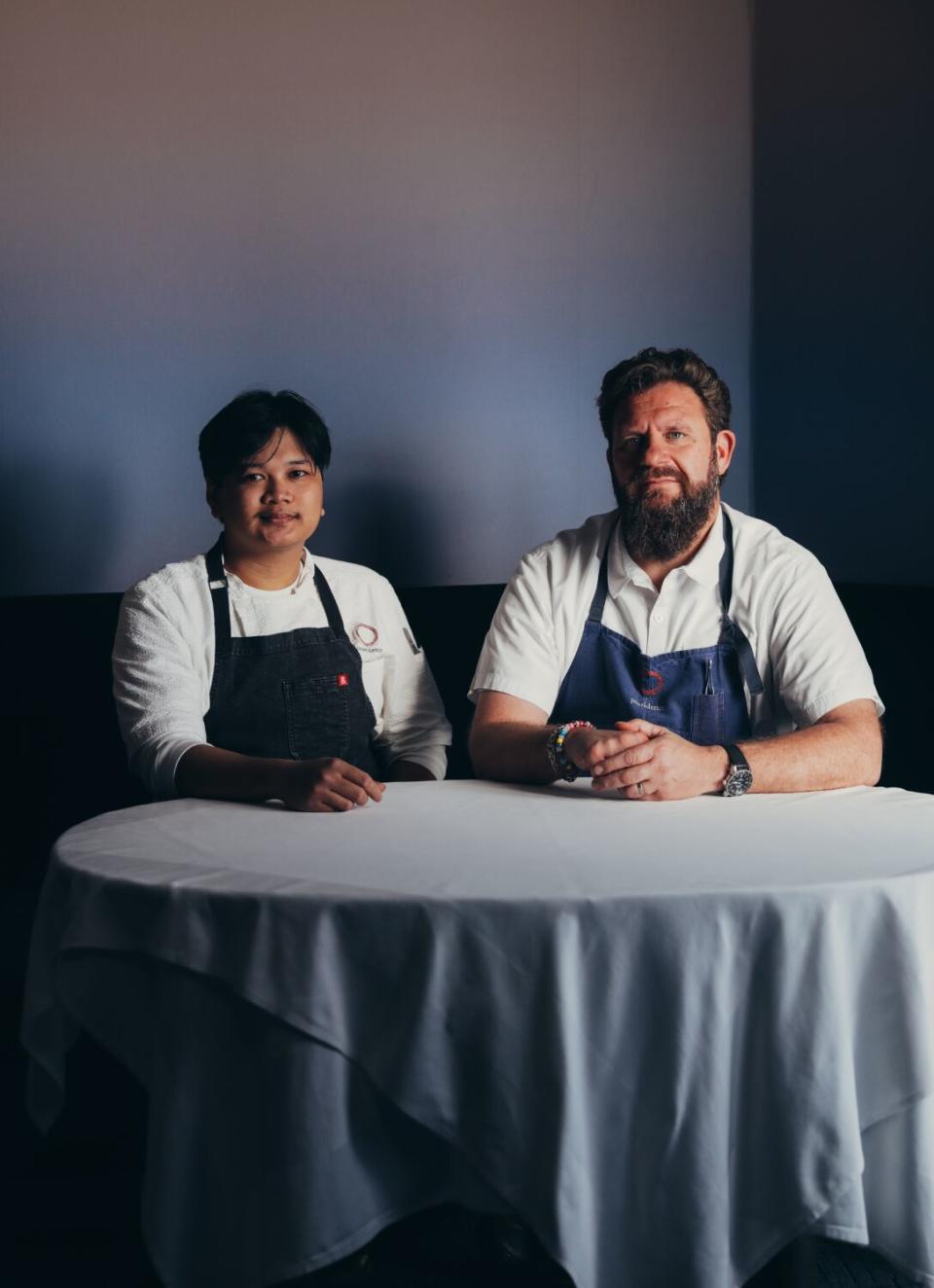 Two men in chef's aprons sit at a table that's covered in a long tablecloth.