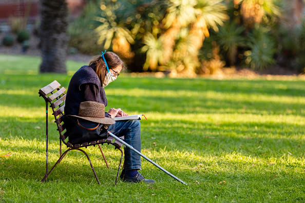 A woman wearing a hand made face mask enjoys the freedom of legally sitting on a park bench to read a book at the Botanical Gardens as restrictions are being eased in Victoria.