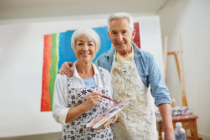 Two smiling people, one holding an artist's palette, with painting behind them.