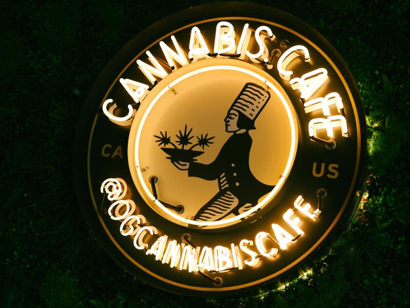 A circular neon sign with the words Cannabis Cafe and @OGCannabiscafe against a leafy green wall
