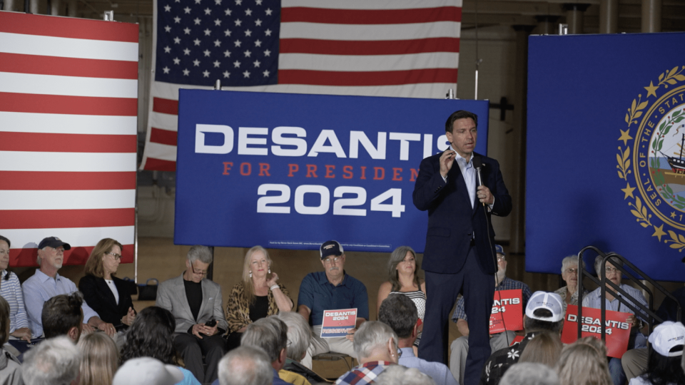 Ron DeSantis speaks at a Never Back Down-hosted event, with super PAC signage and logos behind him. (Alec Hernández / NBC News)