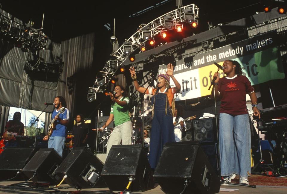 one man and three women sing into microphones on a large stage, the man also plays electric guitar