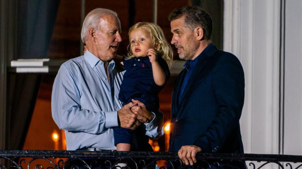 joe biden, wearing a blue dress shirt, holds his young grandson beau while standing next to hunter biden, who wears a dark blue suit jacket, as they stand in front of a railing