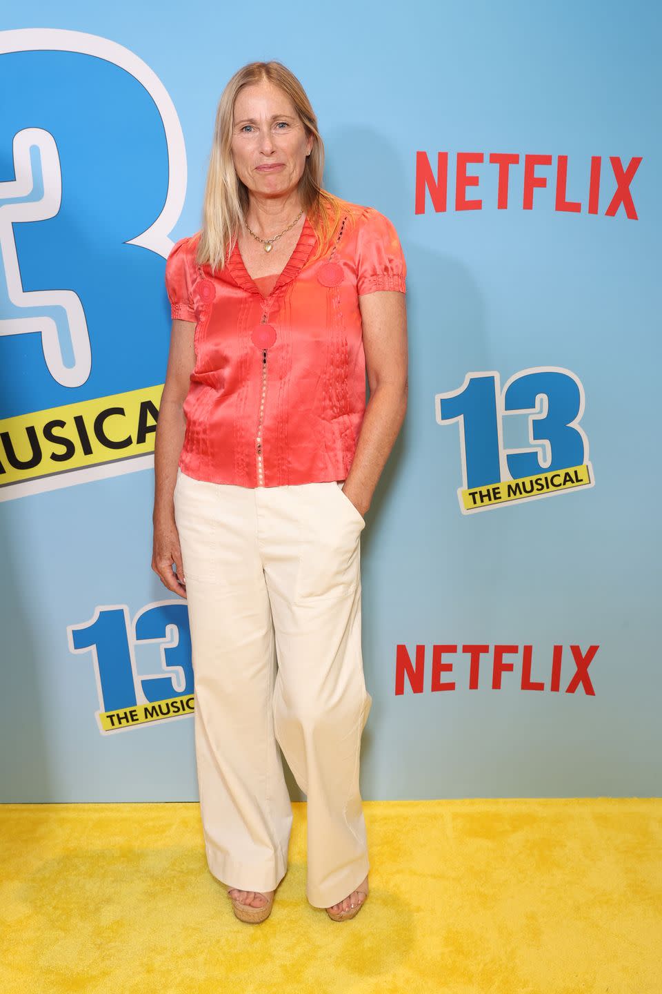 tamra davis wears a coral, short sleeved blouse and cream trousers with peep toe platform heels, she has one hand in her pocket