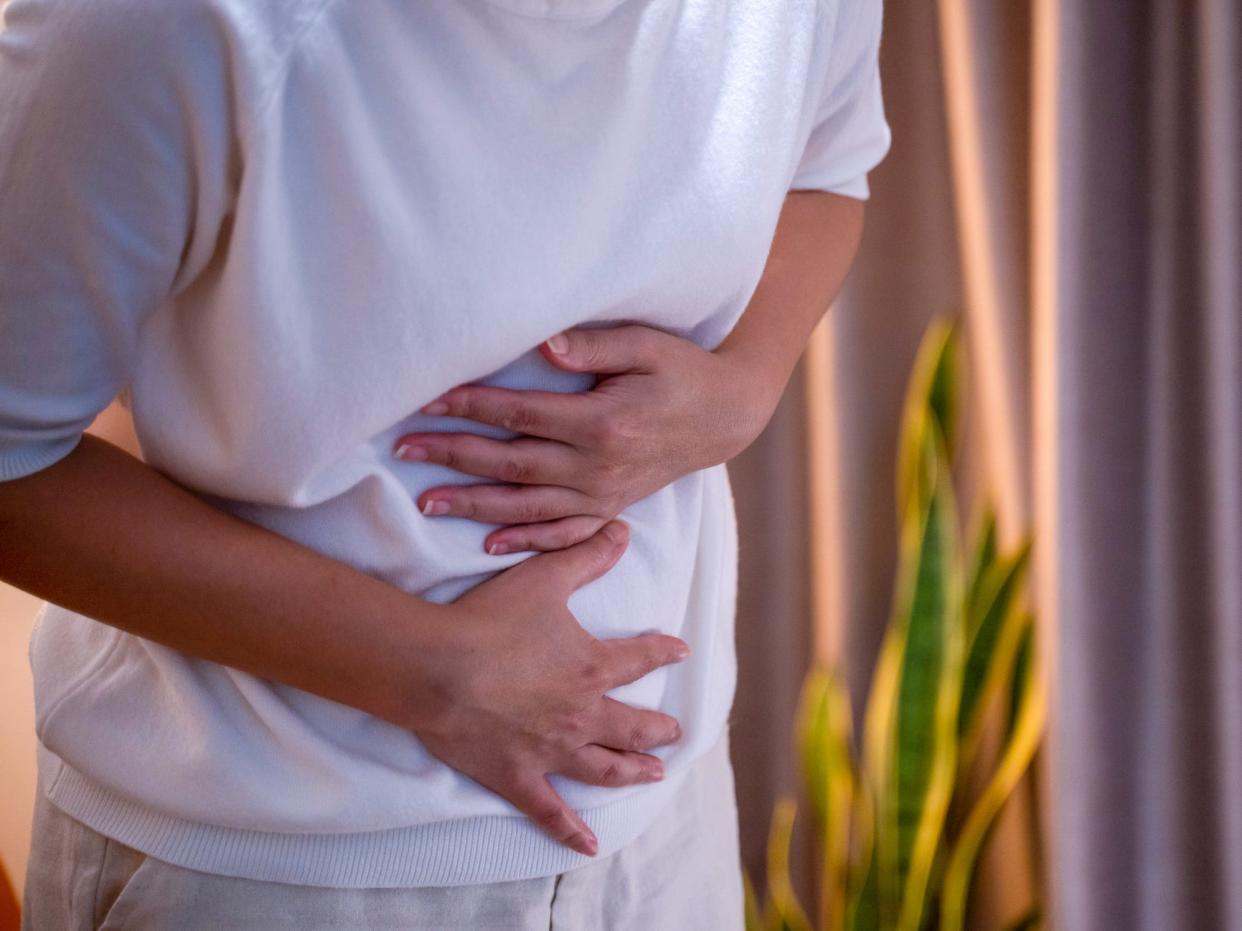 Image of the shoulders down of a person clutching their stomach. The person wears a white sweatshirt and sweatpants and stands on the left side of the viewer, turned to the viewer's right. In the background, you can see a bright green snake plant and pink curtains.