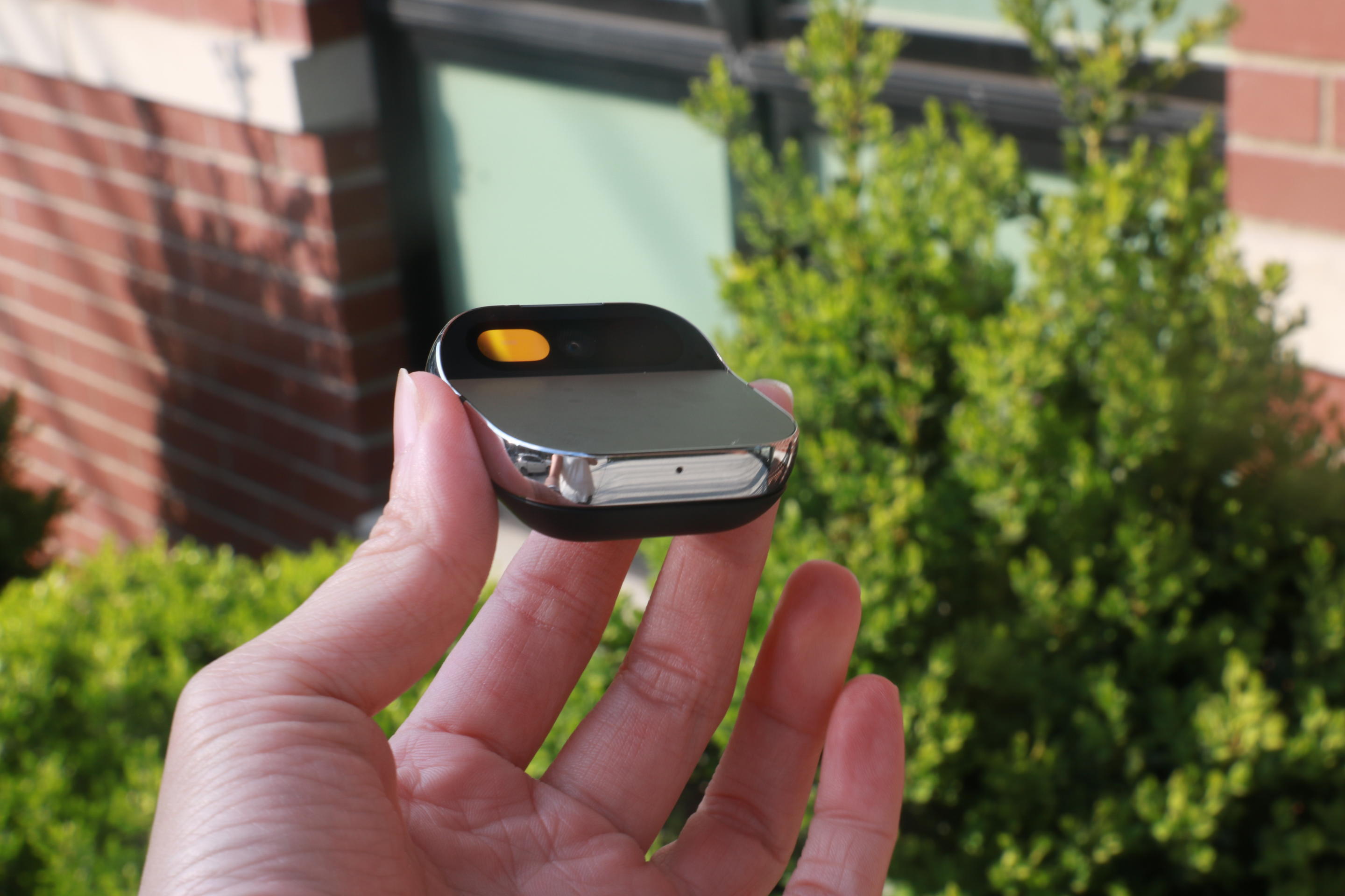 Side view of the Humane AI Pin held in mid-air in front of some green foliage and a red brick building.