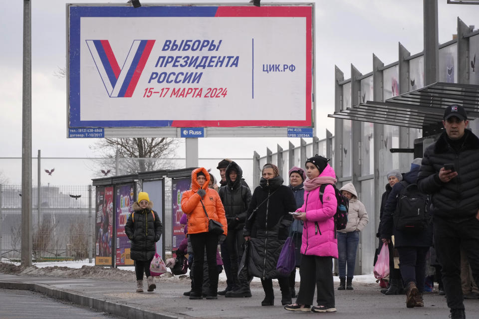 Varias personas esperan en una parada de autobús junto a un anuncio que promueve las próximas elecciones presidenciales en Rusia, en San Petersburgo, Rusia, el miércoles 20 de diciembre de 203. (AP Foto/Dmitri Lovetsky)
