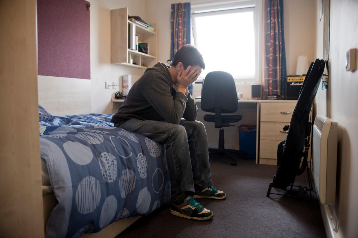 Young Caucasian male student with head in hands, sitting on a bed, alone in room away from home