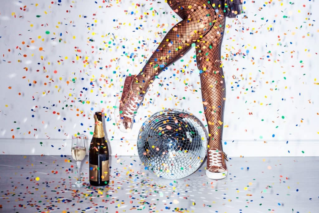 A disco ball sits on the floor with Champagne and confetti in the air. A woman wearing sneakers and a dress is also shown.