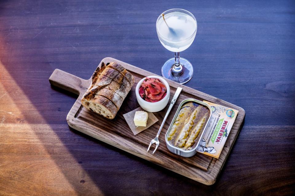 A wood with canned fish, bread, tomatoes and butter next to a glass