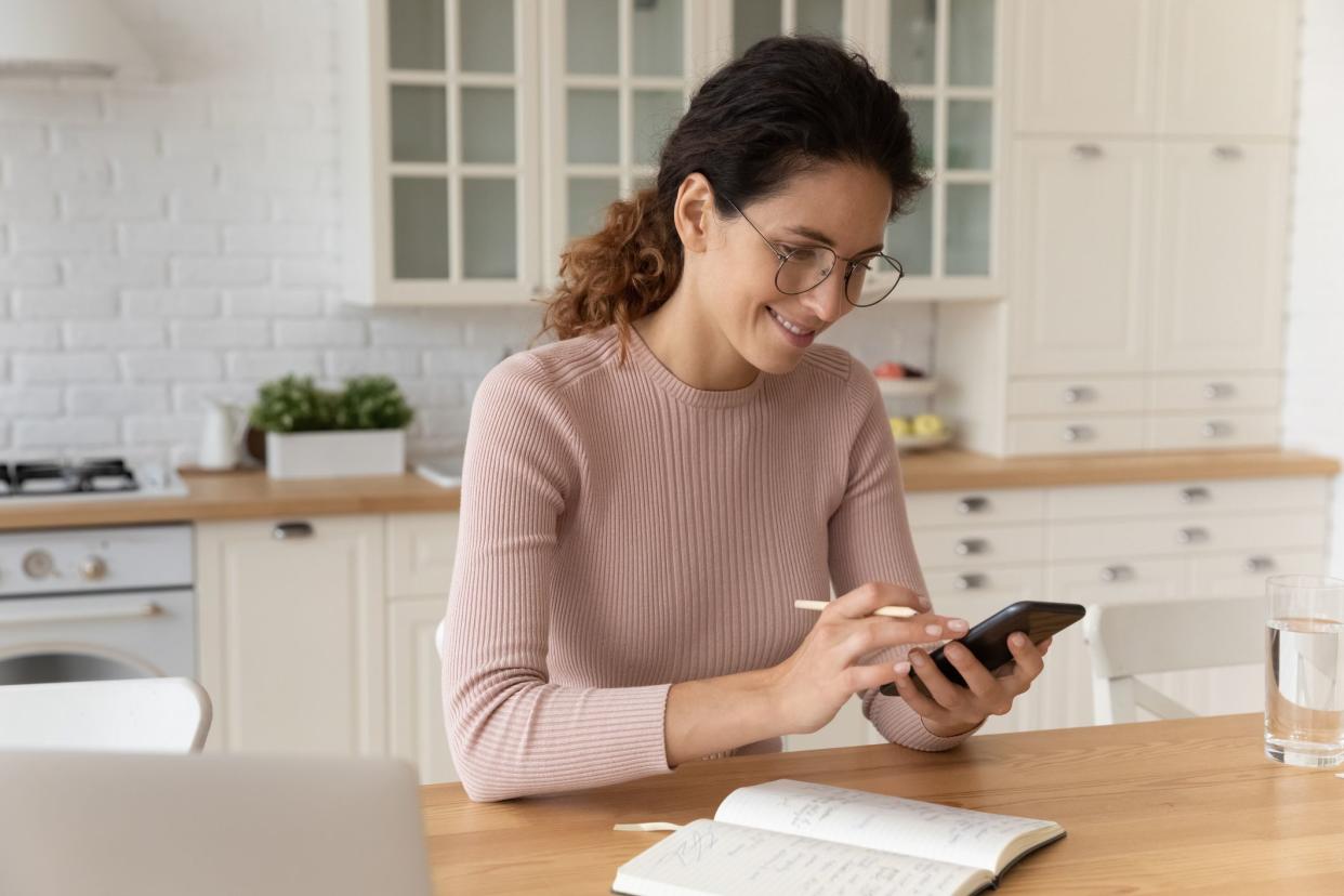 Young hispanic woman in eyeglasses sit at dining table holding smartphone and pencil, check schedule online agenda plan household, surf information on internet writes it in diary, modern tech concept