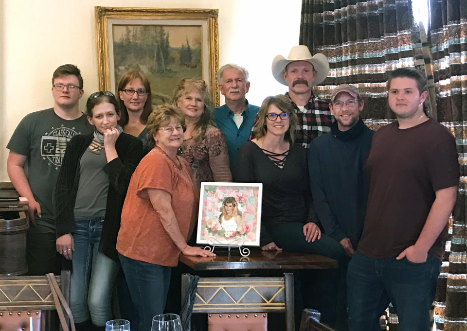 This photo taken March 1, 2021 and provided by Yavapai County prosecutor Susan Eazer shows members of the Pitts family surrounding a photo of Pamela Pitts who was killed in 1988 outside Prescott, Ariz. Pitts then-roommate, Shelly Harmon, confessed to the killing in March 2021 after already having served time for the 1991 death of her ex-boyfriend. From left, Rhys Tarver, Becky Pitts, Tena Pitts, Carol Pitts (orange shirt), Sara Pitts, Paul Pitts Sr., Kerrie Tarver, Jarrett Tarver, Paul Pitts Jr. and Brett Tarver. (Susan Eazer via AP)