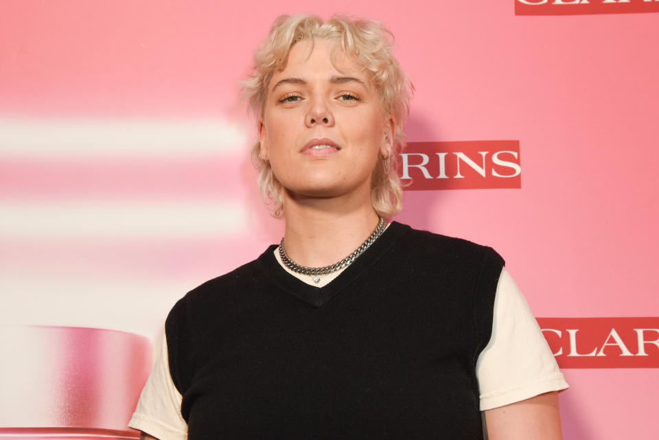 Person on a red carpet wearing a sleeveless black top over a white shirt, posing in front of a Clarins backdrop